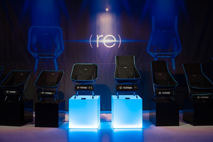 Lineup of black Helinox camp chairs with blue trim displayed on pedestals in front of a (re) backdrop