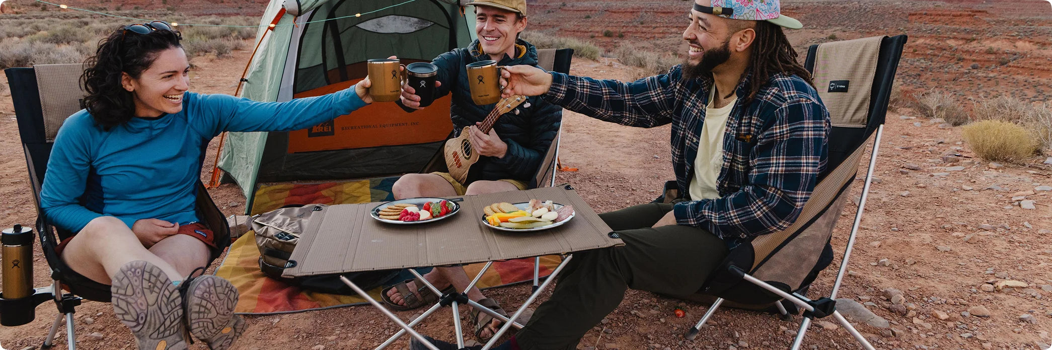 Three friends toasting Hydro Flask mugs around a Helinox table