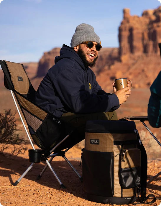 Camper relaxing with a mug beside a Helinox x Osprey cooler